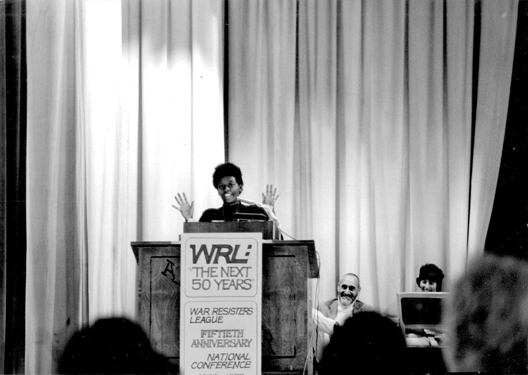 Mandy at the WRL Anniversary Conference podium with Ira Sandperl and Irma Zigas in the background on right; August 7, 1973; Asilomar Conference Center, Pacific Grove, CA. Photo Credit: Nancy Purser. Mandy shares, “I joined the staff of the War Resisters League’s Western Regional Office in San Francisco in 1969. It was my first-ever paying job. I was one of the conference coordinators.” Photo courtesy of Mandy Carter.