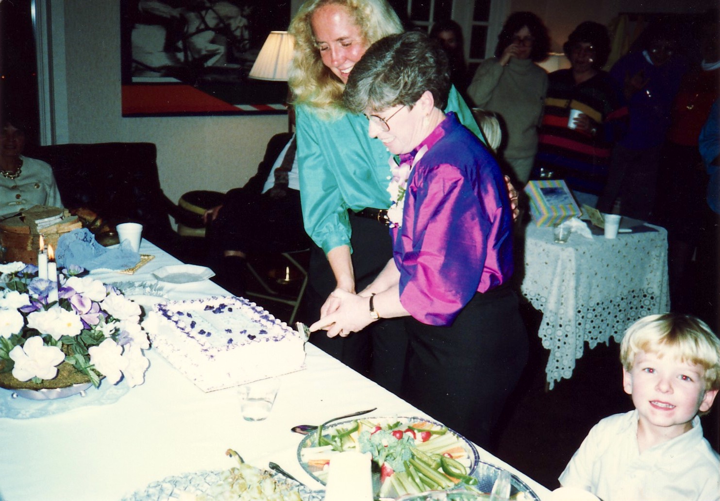 Jennifer Crossen and Joan Callahan (her partner) cut into a cake to celebrate their commitment ceremony while their son David Crossen smiles at the camera, Lexington, KY, 1989. The ceremony was officiated at their house with a minister from the Unitarian Church.