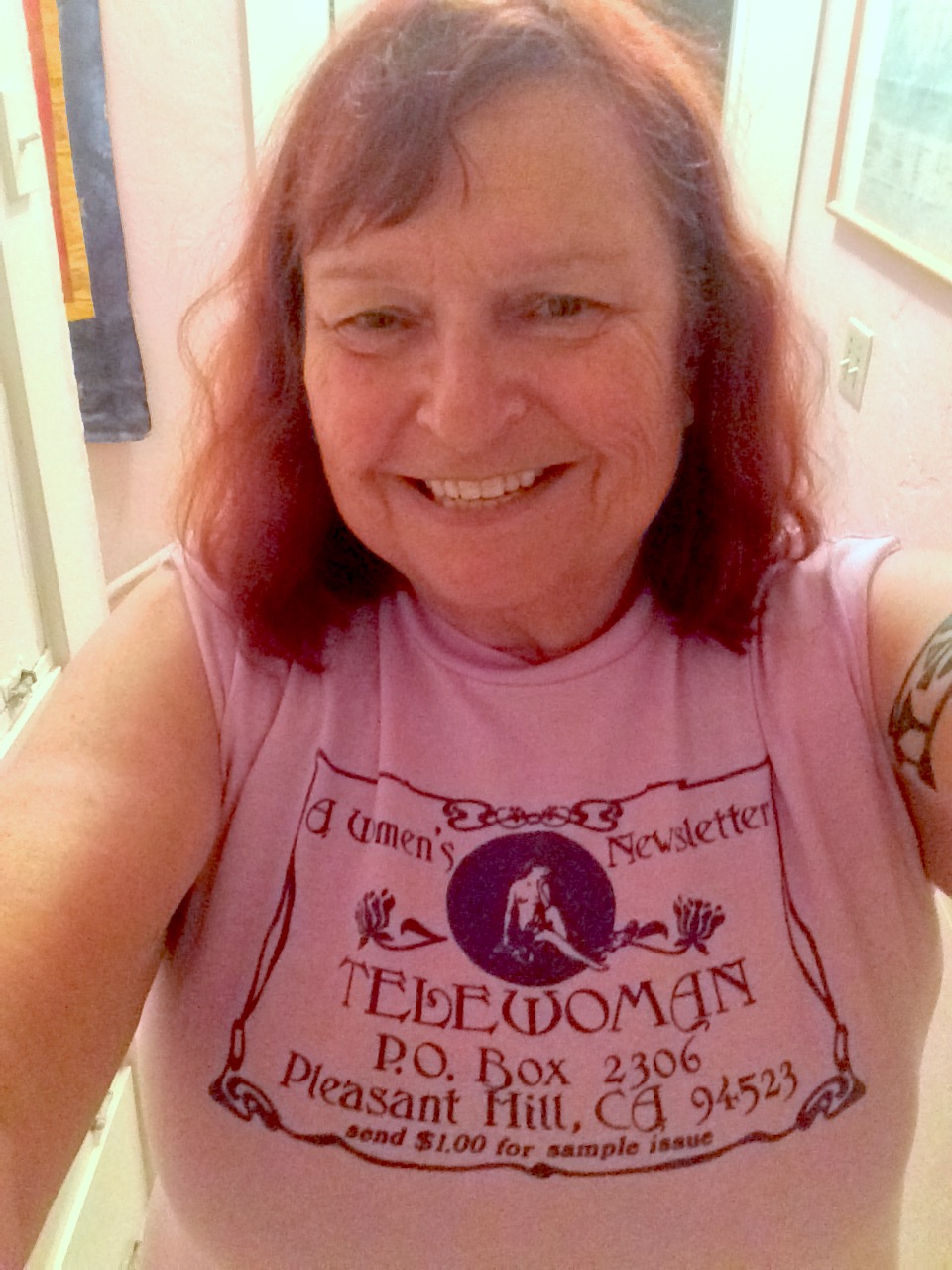 Beth wearing a light pink shirt that reads “Telewoman: A Women’s Newsletter”, 2016. Beth shares,	“In 1983, Anne D’Arcy, editor of Telewoman, a monthly lesbian literary and networking magazine, dragged my by the scruff of my neck off the lesbian community blacklist. I’d write a column and reviews for Telewoman for three years.” Photo courtesy of Beth Elliott.