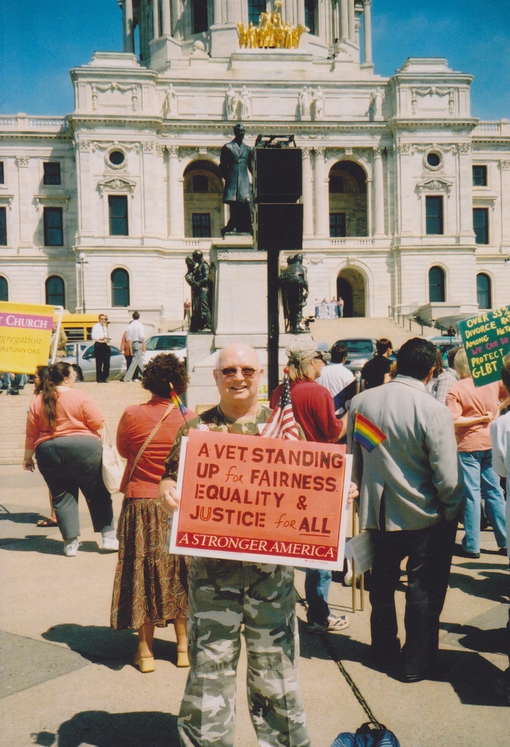 Don Quaintance during Lobby Day at the State Capitol, St. Paul, MN, 2006. He wears military fatigues while holding an American flag and a sign that reads, “A Vet Standing Up for Fairness, Equality, and Justice for ALL.” The sign’s border says, “A Stronger America.” Photo courtesy of Don Quaintance. 
