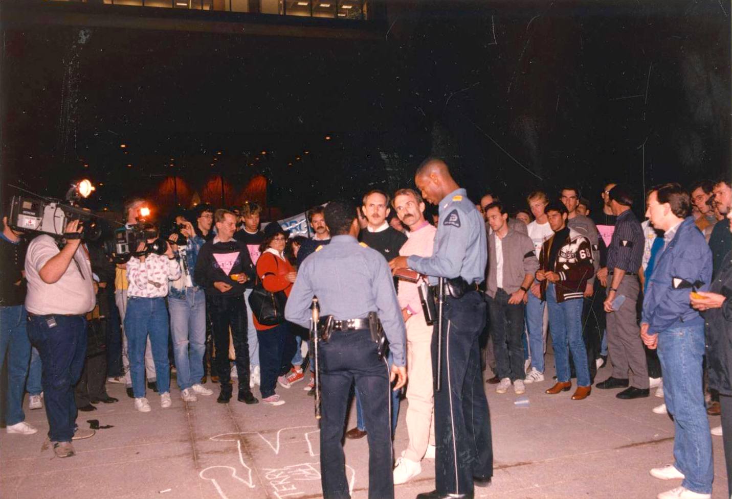 Bill Nelson and William Waybourn speaking with two police officers at the City Hall Plaza memorial for Terry Tebedo after his funeral earlier that day, Dallas, TX, January 28th, 1988. They are standing next to a chalk outline of a body on the sidewalk that represents Nelson's partner, Terry Tebedo. Written inside the chalk outline is the number 641, representing the number of AIDS casualties at the time of Tebedo’s death in 1988. Photo courtesy of William Waybourn.