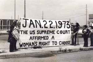 Susan Allen on the far left (with a sign reading “Mandatory Motherhood is UN-AMERICAN”) examining a banner she made for a demonstration against the Anti-Choice Conference, Metairie, LA, 1979.