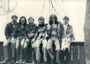 Six white men on a fence, Tom Mosmiller pictured as the 4th man to the right, 1979. These men were members of the nationwide collective in 1979 that launched, produced and published M: gentle men for gender justice which became Changing Men: Issues in Sex, Gender and Politics in 1985.