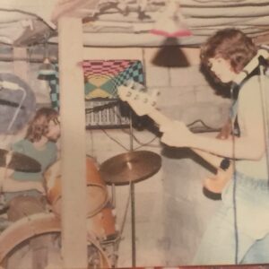 Chris Freeman playing with Jack (drummer) for his first band Relic in the basement of his house, Lake City in Seattle, WA, 1979. Photo credit: Erik Aandal. Chris shares, “The ceiling was so low, I could not stand up all the way. Note the psychedelic poster, Peavey PA head and homemade strobe using a styrofoam wheel with a hole in it on a fan motor.” Photo courtesy of Chris Freeman.
