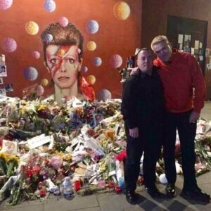 Mehmet and his partner Sepe Vaisanen hugging in front of the David Bowie mural in Brixton, South London. Multiple bouquets of flowers, candles, photos, and notes are laid on the ground around the mural or taped to it. Photo courtesy of Mehmet Sander.