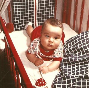 A portrait of Mehmet as a baby, lying in a red crib next to a dotted rattle. Photo courtesy of Mehmet Sander.