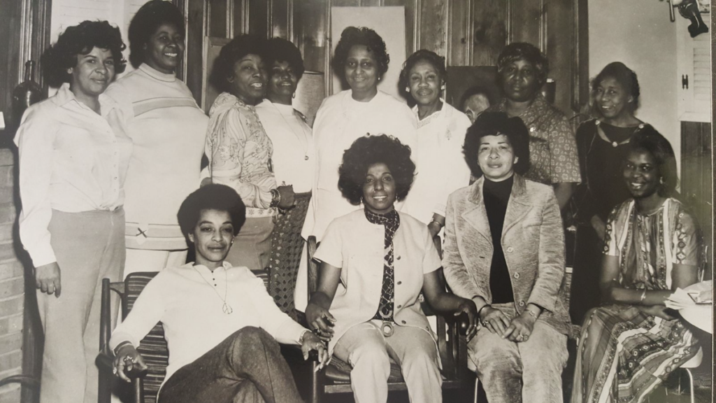 Black and white photo of a meeting of the National Black Nurses Association. Among the group, a young Phyllis Jenkins sits in a low chair, wearing a white henley shirt, peace sign necklace, and a short Afro.