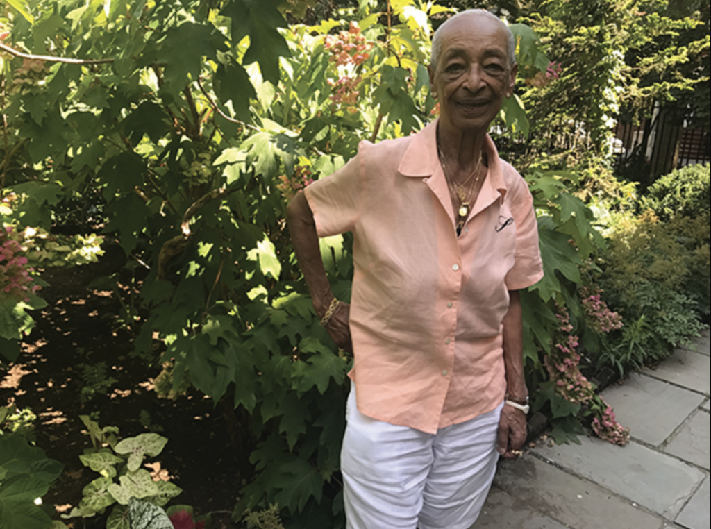 Phyllis Jenkins, a Black woman in her 80s, stands in a garden smiling, with her right hand on her hip. She wears a peach blouse, white slacks, and gold jewelry.