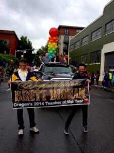 Brett riding in the Portland Pride Parade, Portland, OR, 2014. The banner ahead of him reads, “Congratulations Brett Bigham, Oregon’s 2014 Teacher of the Year”. Photo courtesy of Brett Bigham.