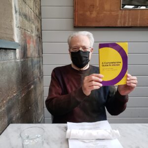 Father Tom Picton holding a purchased copy of Jay’s book “A Comprehensive Guide to Intersex” and wearing a mask to protect against COVID-19 in Seattle, WA. Jay shares, “Father Picton is my Tom Picton, my priest, confidant, and friend. He listened to me read and think through pages of the manuscript of my book for countless hours. He has been my recovery support person for over 8 years.” Photo courtesy of Jay Kyle Petersen.