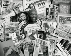 Rupert underneath a pile of publications, depicting his career as an Art Director, Cartoonist, and freelance graphic designer for various publications, theater groups, and progressive and grassroots organizations, 1992.