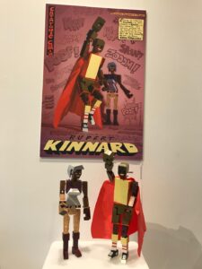 Block action figures of Rupert’s characters B.B. and the Diva, placed in front of a related poster, at the 2019 Portland Design for Good Initiative’s “Staying Out: Pioneers of Pride” exhibit. Rupert shares, “New York artist Chris Chan was selected to honor Cathartic Comics.”