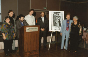 L-R: Judy Abdo, Dennis Zane (former Santa Monica City Council Member), Ken Genser (speaking at podium; former Santa Monica City Council Member and Mayor), Unidentified, and Tony Vasquez (former Mayor, Santa Monica City Council Member, and State Board of Equalization Member) at Herman and Millie Rosenstein’s wedding anniversary celebration. A signed card featuring an early portrait of the Rosensteins is featured at left of the couple, who are smiling and looking on from the righthand side. Photo courtesy of Santa Monica Public Library Image Archives, Judy Abdo Photo Album Collection.