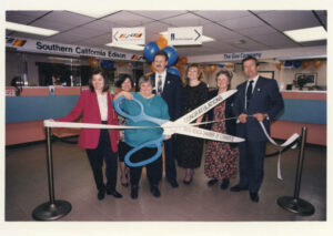 Judy Abdo cutting through a white ribbon, which is held by others on the right and left sides, to officially open the Southern California Edison Office in Santa Monica, CA, 1990s. The scissors read, “Congratulations! Santa Monica Chamber of Commerce”. Photo courtesy of Santa Monica Public Library Image Archives, Judy Abdo Photo Album Collection.
