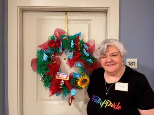 Deborah smiling outside the door of her apartment at Law Harrington Senior Living Center, which was was by an LGBTQA+ organization called the Montrose Center. Her shirt reads, “Full of Pride” in rainbow lettering. The wreath reads, “There’s no place like home!” with Wizard of Oz characters placed around it. Photo courtesy of Deborah Moncrief Bell.