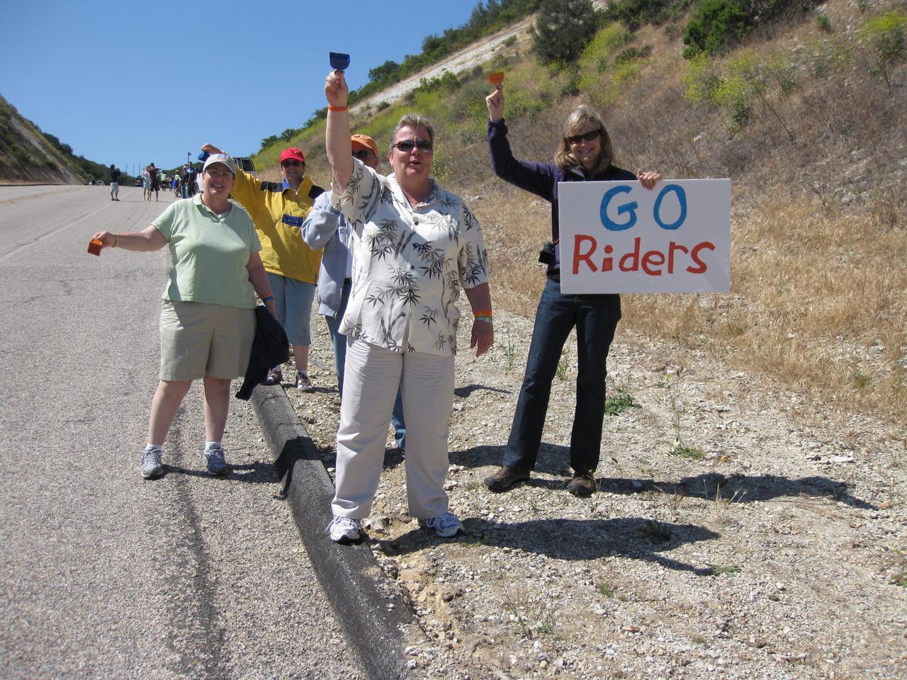 Lorri and her wife Gina Calvelli (on left) cheering on AIDS/LifeCycle riders at the top of the “Evil Twins”. Another participant holds a sign that reads, “Go Riders”.
