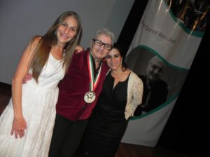 L-R: Sarah Mondragón (daughter), Margaret Randall, and Ximena Mondragón (daughter) smiling at the award ceremony for Margaret’s Medal for Literary Merit in Ciudad Juárez, Chihuahua, Mexico, 2017. The awarding organization was Literatura en el Bravo, and the banner behind them features a portrait of Margaret. Photo courtesy of Margaret Randall.