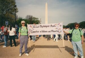 Esther Rothblum and Marcia Hill participating in the March on Washington for Lesbian, Gay, and Bi Equal Rights and Liberation on the National Mall, Washington, DC, April 25, 1993. They wear lime green “Committee on Women and Minorities” t-shirts and hold a sign that reads, “Vermont Psychological Association Committee on Women and Minorities” with flowers along the edges. Photo courtesy of Esther Rothblum.