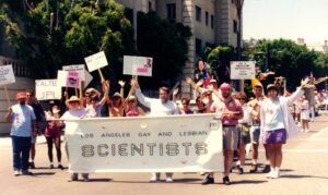 Over a dozen members from the Los Angeles Gay and Lesbian Scientists marching in the West Hollywood Pride Festival and Parade, West Hollywood, CA, June 1992. The group won the Grand Marshal's Award for their Pride=Power entry and marched with white and pink-colored signs behind a white banner that reads, “Los Angeles Gay and Lesbian Scientists”. 