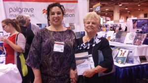 Dr. Andrea L. Dottolo (at left) and Dr. Oliva Espin (at right) smiling in front of the Palgrave-Macmillian booth during the American Psychological Association conference. Oliva holds a copy of “Gendered Journeys: Women, Migration, and Feminist Psychology” (2015), which they co-authored. Photo courtesy of Oliva Espín.