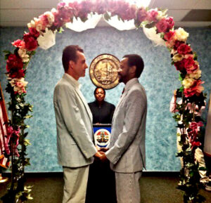 Paul Outlaw (at right) with partner Ray Busmann (at left) facing each other under a flower-arch while getting legally married at the Los Angeles County Registrar-Recorder/County Clerk’s office, Norwalk, CA, July 17, 2013. Photo courtesy of Paul Outlaw.
