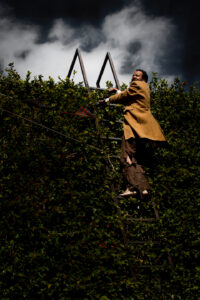 Paul performing in the world premiere of his play The Late, Late Show at Bootleg Theater in Los Angeles, CA, 2013. He's outdoors, wearing a Victorian topcoat and frayed trousers, climbing up a ladder wedged against a high wall covered in ivy. Photo credit: Scott Pitts. Photo courtesy of Paul Outlaw.