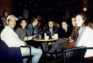 Dante Alencastre (Left) in a baseball cap smiles while sitting with a group of film festival 
friends on a bar patio where he screened his 1st narrative short, 