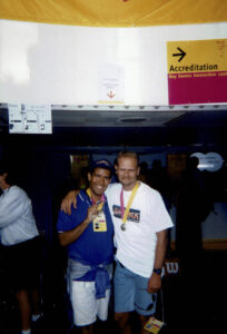 Dante and Ron smile, arms around each other and holding up medals under a sign for the Gay Games in Amsterdam, 1998; during which they both competed as swimmers. Photo courtesy of Dante Alencastre. 