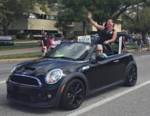 Lori Haas drives Stephanie (backseat, standing) joyfully waves to passerby in her beloved Mini Cooper Roadster Convertible in the Wichita Pride Parade, Kansas, 2018. Photo courtesy of Stephanie Byers.