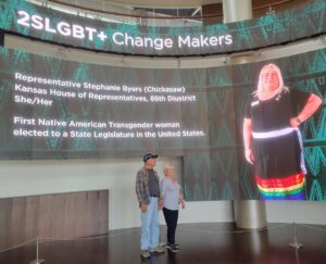 Photo of Stephanie Byers’ parents looking at a display of “2SLGBT+ Change Makers” with a large photo of Stephanie herself, in the First Americans Museum in Oklahoma City, Oklahoma, 2022. Photo courtesy of Stephanie Byers.