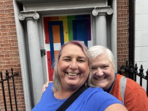 Stephanie Byers smiles and hugs with her wife Lori Haas outside a rainbow painted front door in Dublin, Ireland, 2022. Photo courtesy of Stephanie Byers.