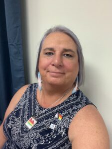 Stephanie smiles in trans-flag colored beaded earrings, while at a speaking event at Kansas City Community College in Kansas City, Kansas, 2023. Photo courtesy of Stephanie Byers.