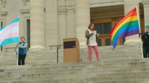 Stephanie Byers, flanked by the Trans and Rainbow Pride flags, speaks at a podium on the steps of the Kansas State Capitol, in Topeka, Kansas, 2017. Photo courtesy of Stephanie Byers.