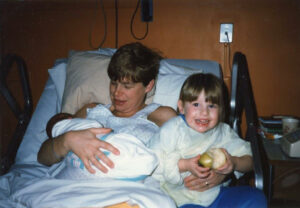 Cidny holding his newborn daughter Jessie Bullens (left) with sister Reid Bullens (age 2, right) smiling at the camera in a medical setting, February 24, 1985. Photo courtesy of Cidny Bullens.