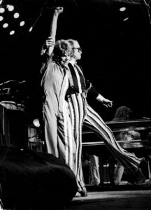 High contrast greyscale photo of Cidny Bullens (L) and Elton John (R) hugging on stage in Foxborough, MA, July 4, 1976. Photo Credit: Mike Hewitson. Photo courtesy of Cidny Bullens.	