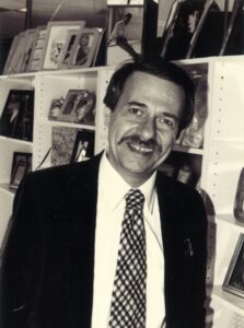 Gerald smiling while standing in front of a wall of personal photos in Denver, Colorado, 1982. Photo courtesy of Gerald Gerash. 