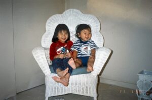 Aiden (left) and younger brother Stefen Aizumi smile and sit in a white rattan chair together. Arcadia, CA, 1992. Photo courtesy of Marsha Aizumi.