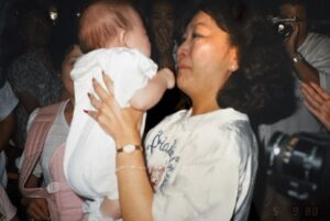 Marsha Aizumi affectionately holds and smiles at newborn baby Aiden for the first time, there are several smiling and photographing/cheering family members in the background dimly lit. LAX, Los Angeles, CA, 1988. Photo courtesy of Marsha Aizumi.