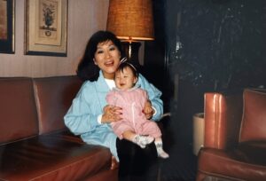 Marsha Aizumi holds baby Aiden (assigned female at birth) on a leather couch, both of them are smiling and laughing together. San Jose, CA, 1989. Photo courtesy of Marsha Aizumi.