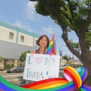 Marsha wears rainbow beads and a Rainbow Pride flag with an “I <3 My Trans Son” sign. Long Beach, CA, circa 2014. Photo courtesy of Marsha Aizumi.