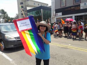 Marsha smiles in a sun hat and holds up a rainbow “Proud PFLAG Mom” flag. San Diego, CA, 2017. Photo courtesy of Marsha Aizumi. 