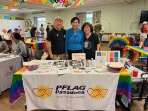 (L-R) Aiden Azumi, U.S. Congresswoman Judy Chu, and Marsha Aizumi smile in front of a PFLAG Pasadena table at a community Pride event in Altadena, CA, circa 2019. Photo courtesy of Marsha Aizumi.