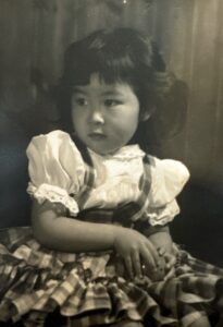 Greyscale photo of Marsha Aizumi as a young girl in a gingham dress looking off camera. Cleveland, OH, circa 1950. Photo courtesy of Marsha Aizumi.