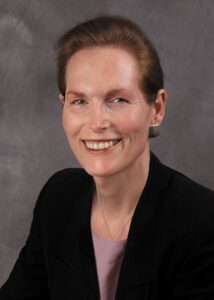 Headshot of Marsha smiling in a black suit and metallic earrings. Photograph courtesy of Marsha Botzer. 