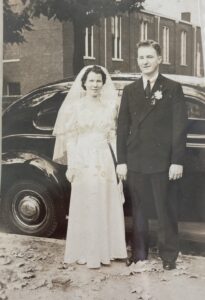 Grayscale Wedding photo of Monica’s parents– Helen and Robert Wehrle smiling in their formal wear in front of a luxury car. In Fort Wayne, Indiana, 1939. Photo Courtesy of Harriet Miller and Monica Wehrle.