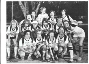 Sepia-toned photo of Harriet Miller and Monica Wehrle with the Fort Wayne Women’s Bureau Softball team joyfully holding up a banner that says “Run, Jane, Run.” In Fort Wayne, Indiana, 1980’s. Photo Courtesy of Harriet Miller and Monica Wehrle.