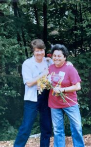 Monica Wehrle and Harriet Miller smile and hug in front of a forest in iconic Lesbos, Greece, 1990. Photo Courtesy of Harriet Miller and Monica Wehrle.