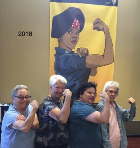 Harriet Miller, Monica Wehrle, Harriet’s daughter Kim, and friend Linda on a historic day 
of Same-Sex Marriage passing baring “Strong flexed arms” in front of a Rosie the Riveter-woman of color banner. In California, 2018. Photo Courtesy of Harriet Miller and Monica Wehrle.