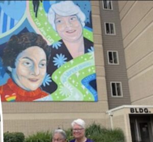 Up-close vibrant photo of Harriet Miller and Monica Wehrle’s public mural, they’re both smiling while standing in the foreground. In Fort Wayne, Indiana, 2023. Photo Courtesy of Harriet Miller and Monica Wehrle.