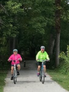 Harriet Miller and Monica Wehrle wear neon pastel outfits while happily riding bicycles side by side on a forest-pathway. In Fort Myers, Florida, 2017. Photo Courtesy of Harriet Miller and Monica Wehrle.